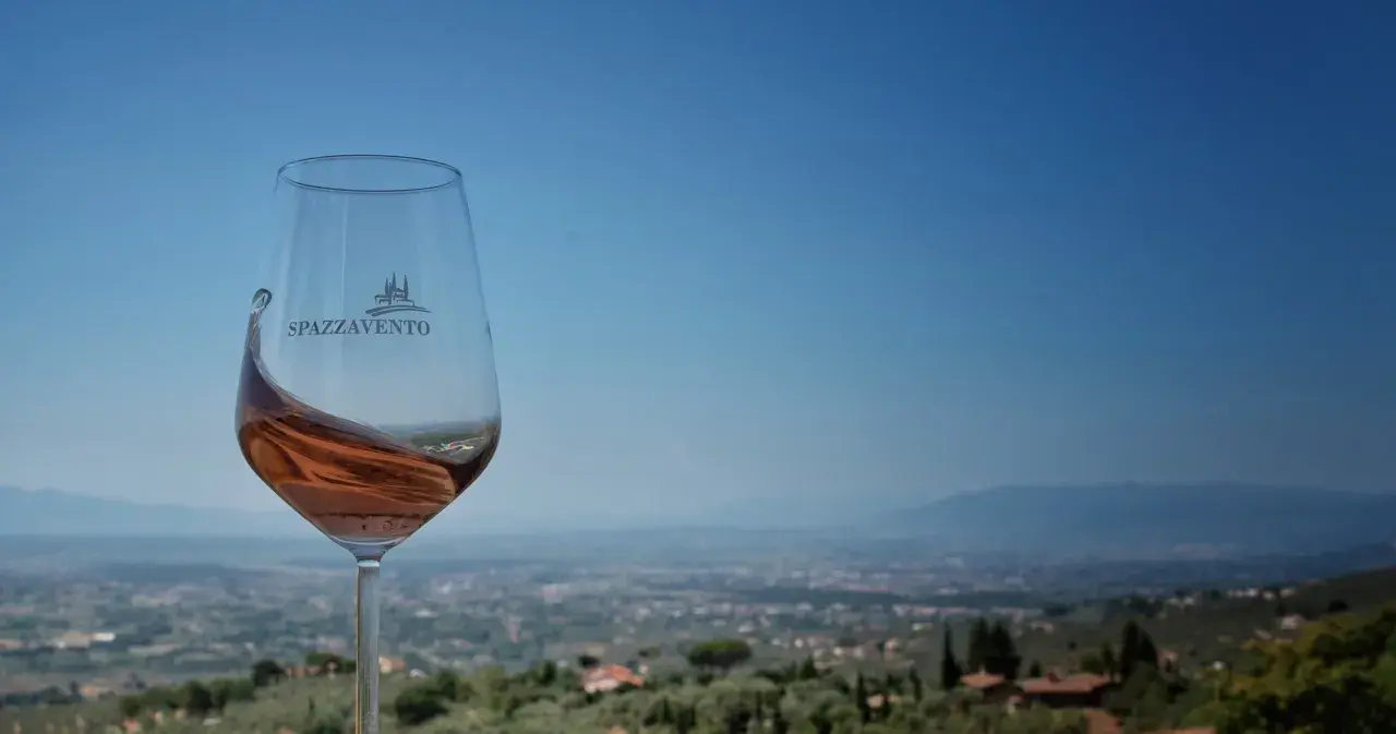 Calice di vino rosato con logo Spazzavento, sullo sfondo il panorama della fattoria di Piazzano e le colline toscane.