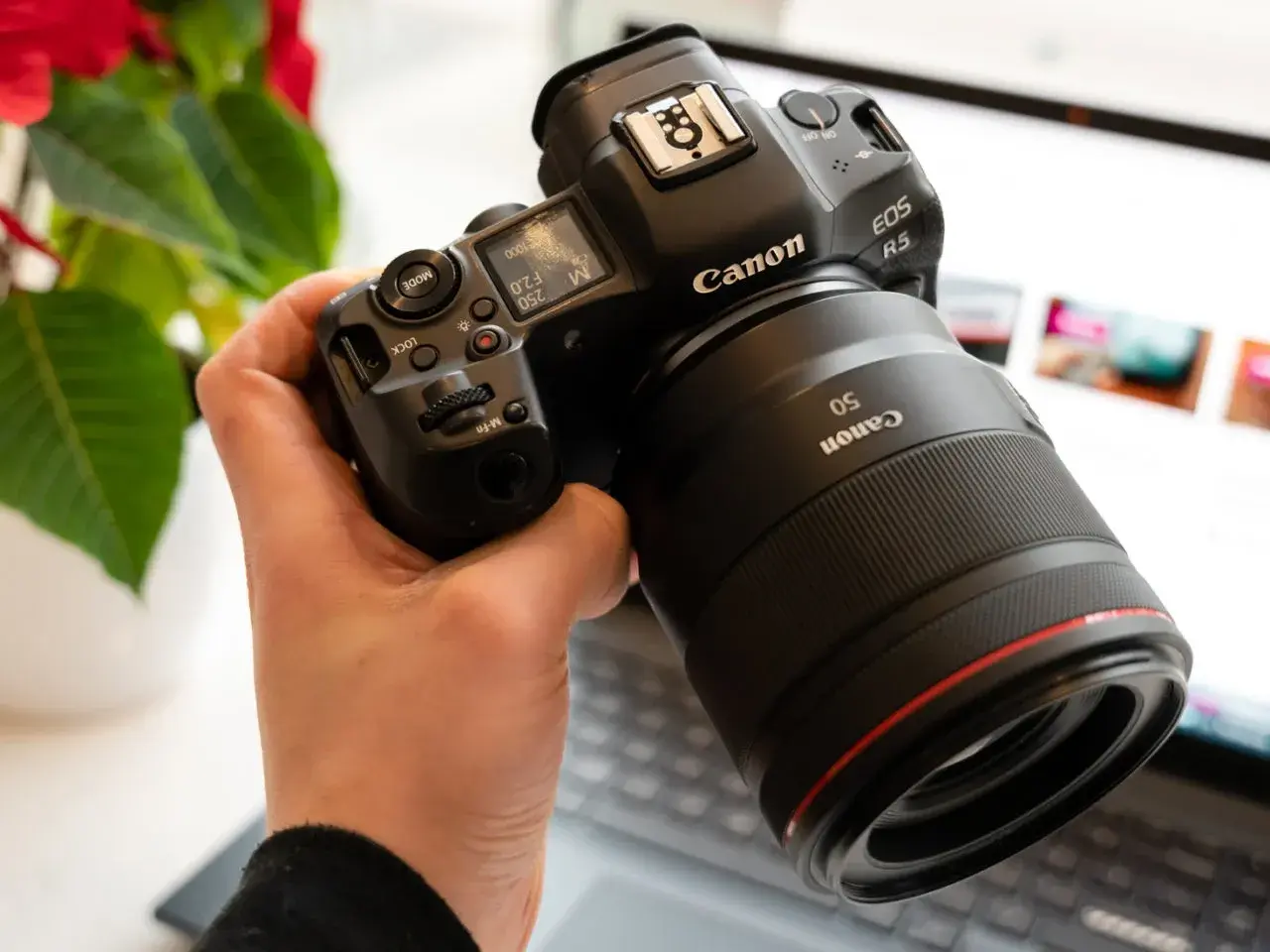 Dłoń trzyma aparat Canon EOS R5 z obiektywem 50mm. W tle widać ekran laptopa.