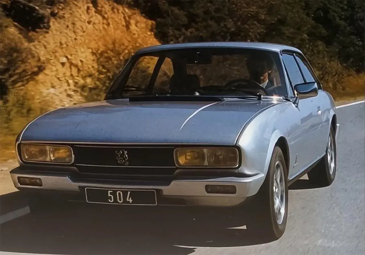 Srebrny Peugeot 504 z lat 70. jedzie po krętej drodze wśród wzgórz.