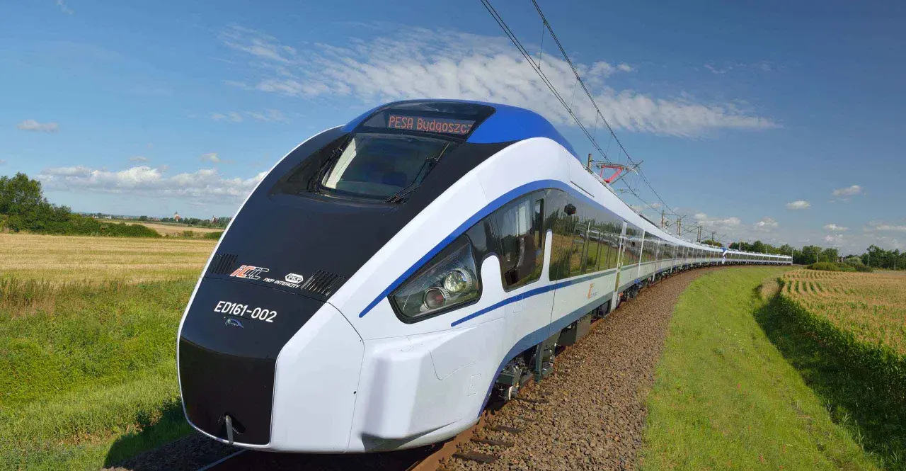 Nowoczesny pociąg Pesa Bydgoszcz ED161-002 mknie po torach wśród pól i zieleni, pod błękitnym niebem z chmurami.