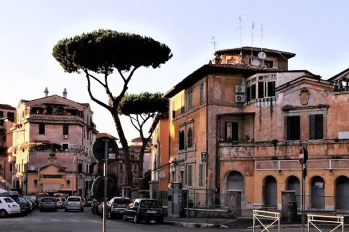 Edifici color ocra e rosa con pini marittimi svettanti, tipici de **la Garbatella**, Roma. Auto parcheggiate lungo la strada.