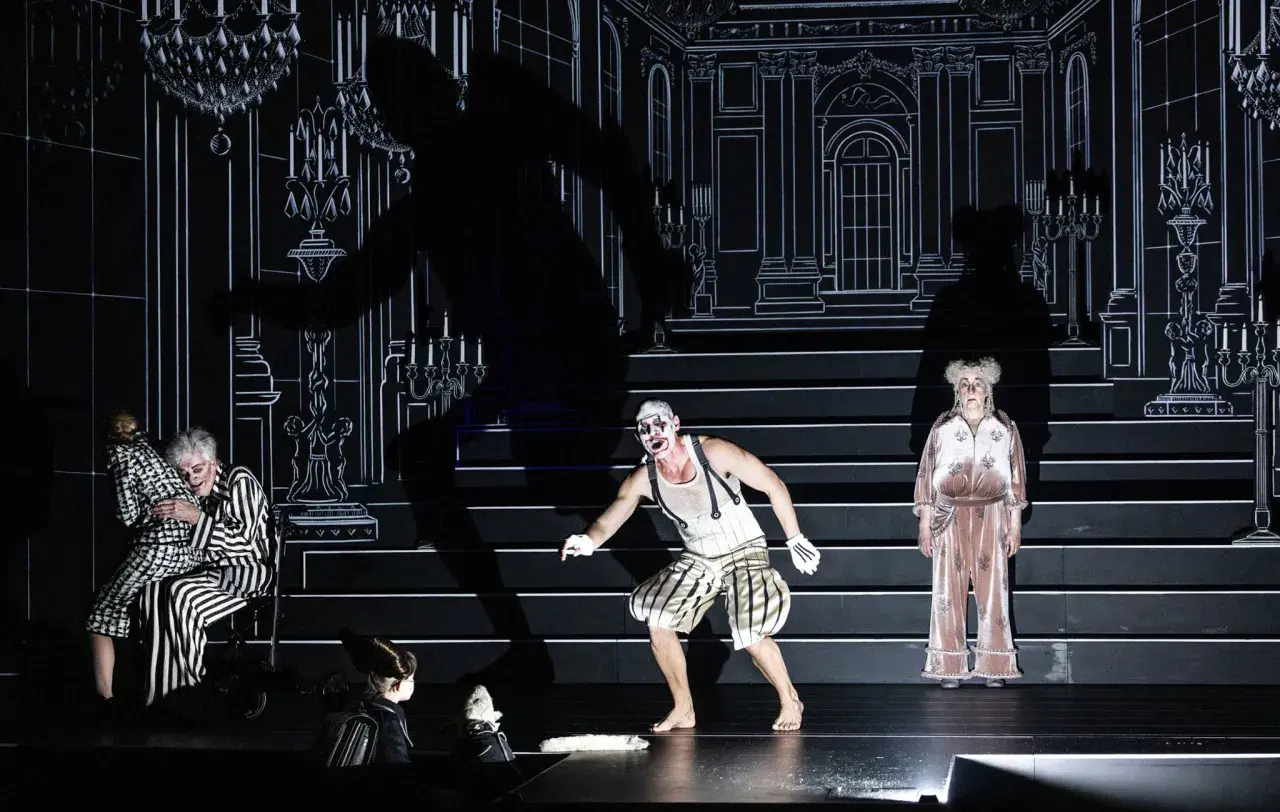 Scen från "Misantropen" på Dramaten. En clownliknande figur, en kvinna i randigt och en äldre kvinna i rosa pyjamas.