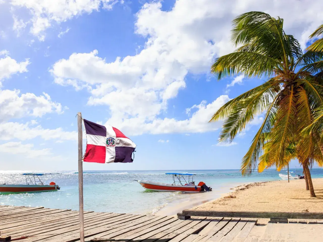 Dominikanische Flagge weht am Strand. Boote im türkisfarbenen Meer. Palmen. Hier ist es im Januar warm.
