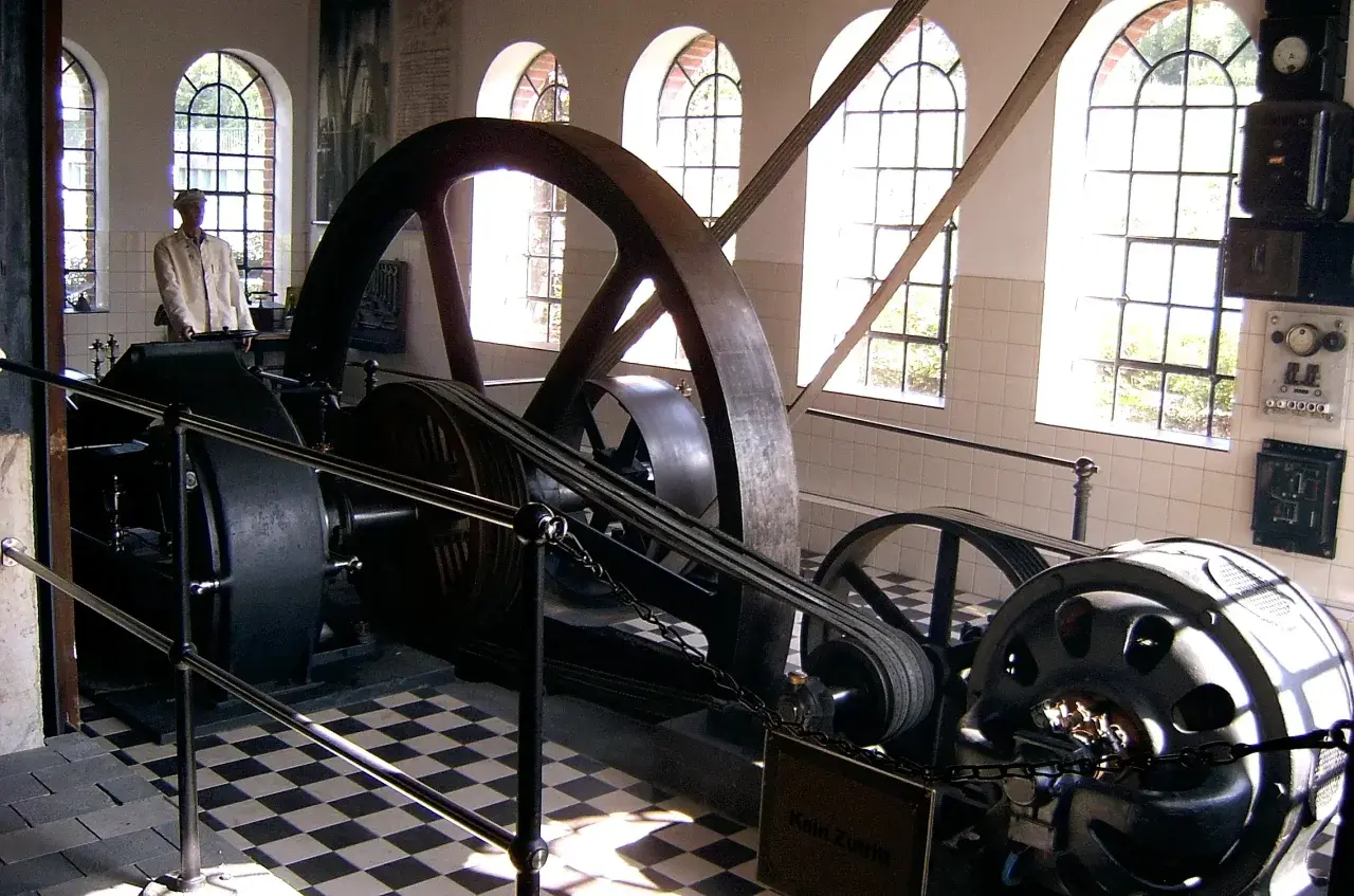 Eine historische Dampfmaschine, Symbol der Industrialisierung, mit großen Schwungrädern und Riemenantrieb. Ein Mann in Arbeitskleidung steht daneben.