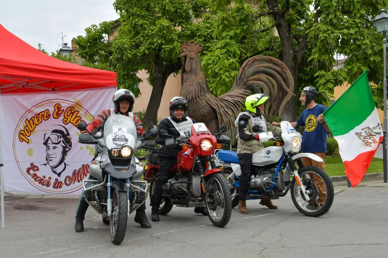 Tre motociclisti pronti per un percorso eroica in moto, con bandiera italiana e un grande gallo di metallo sullo sfondo.