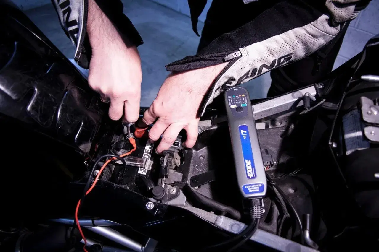 Mani che collegano un caricabatteria Exide alla batteria di una moto. Scopri come caricare batteria moto senza caricabatteria.