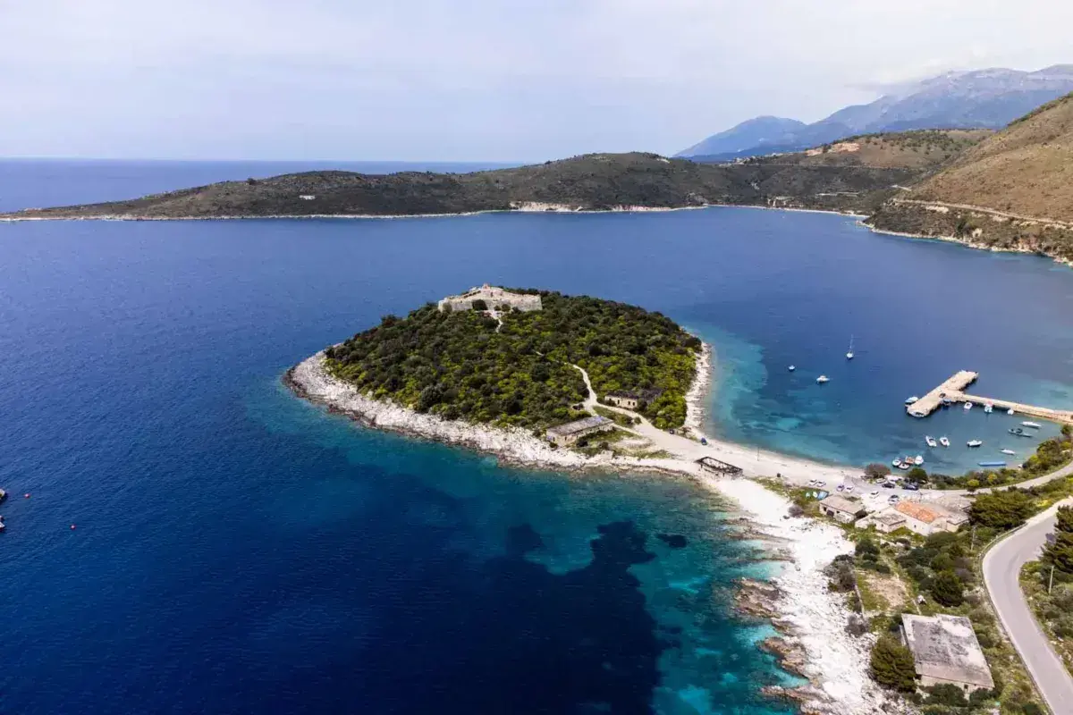 Isla con un castillo en Himare, Albania, rodeada de aguas azules y vegetación exuberante. Un muelle y barcos anclados completan la vista.