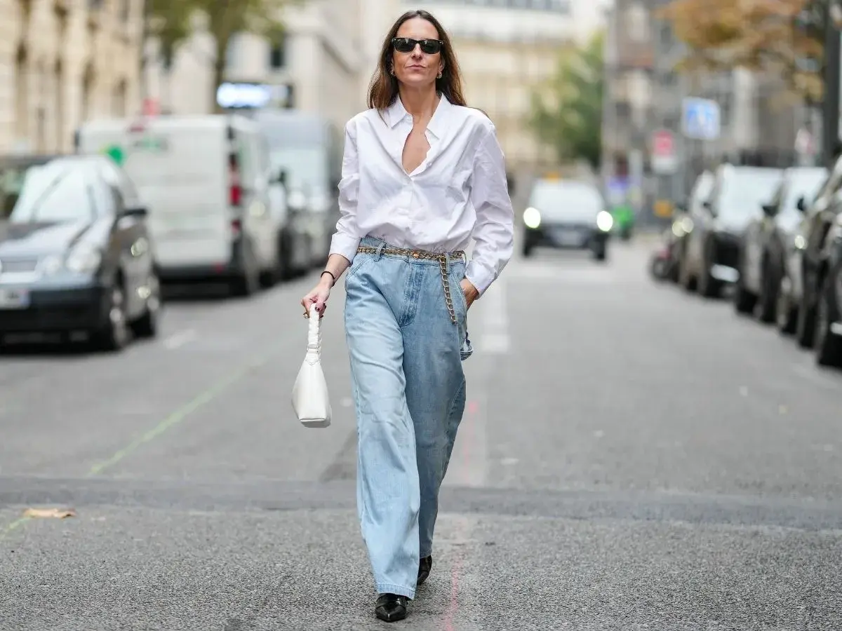 Kobieta w luźnych, jasnych jeansach typu mom fit i białej koszuli, z białą torebką. Stylizacja dla każdej kobiety ceniącej wygodę i modny wygląd.