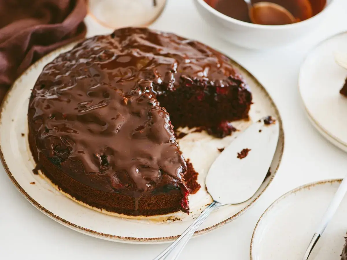 Pyszny brownie z malinami, polane błyszczącą czekoladą, czeka na talerzu. Obok stoi łopatka do ciasta.