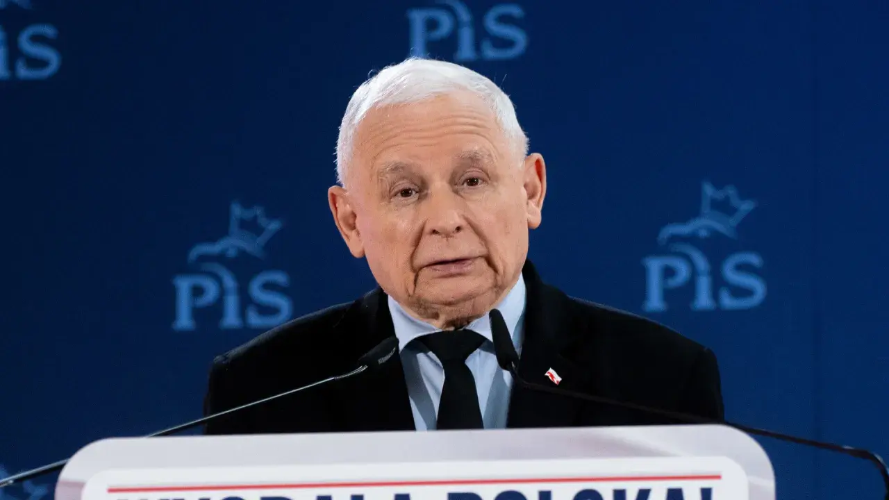 Jarosław Kaczyński w garniturze przemawia za mównicą na tle niebieskiego baneru z logo PiS.