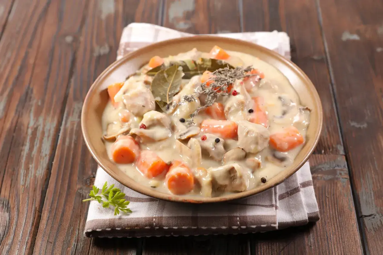 Un plat de blanquette de veau crémeuse avec des carottes et des champignons, garni de thym et de feuilles de laurier.