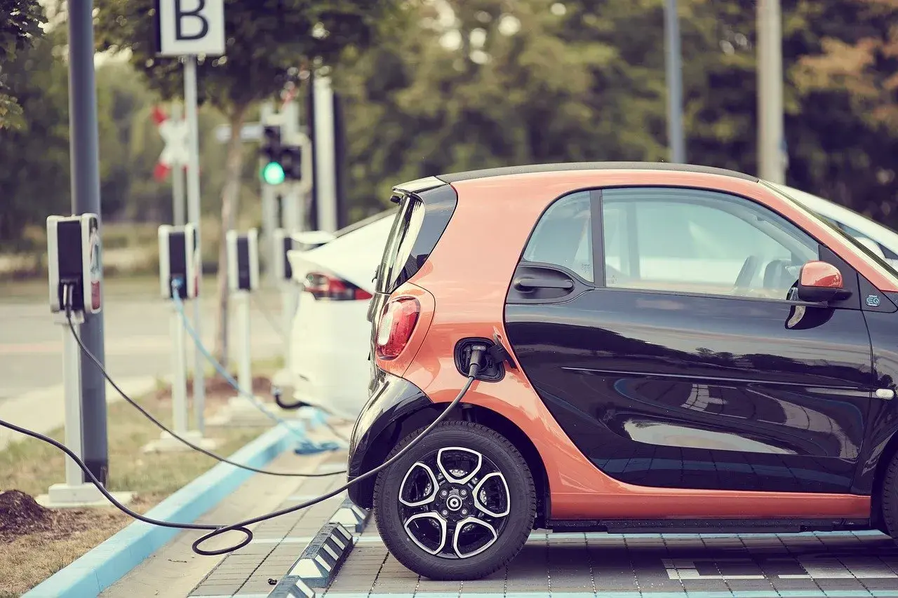 Due **utilitarie elettriche** arancioni e nere, una bianca, sono in carica. Stazioni di ricarica con cavi collegati.
