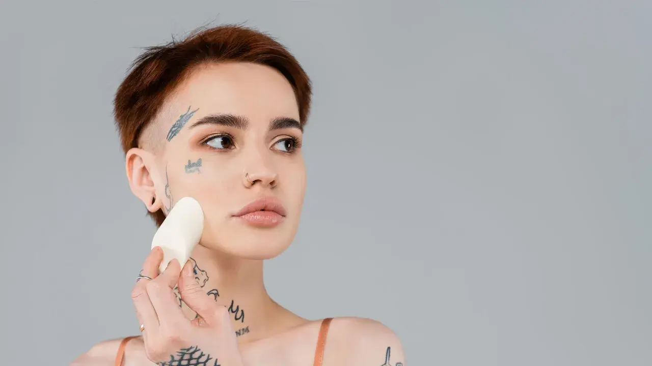 En kvinna med tatueringar applicerar foundation med en svamp för att jämna ut sin underton hud.