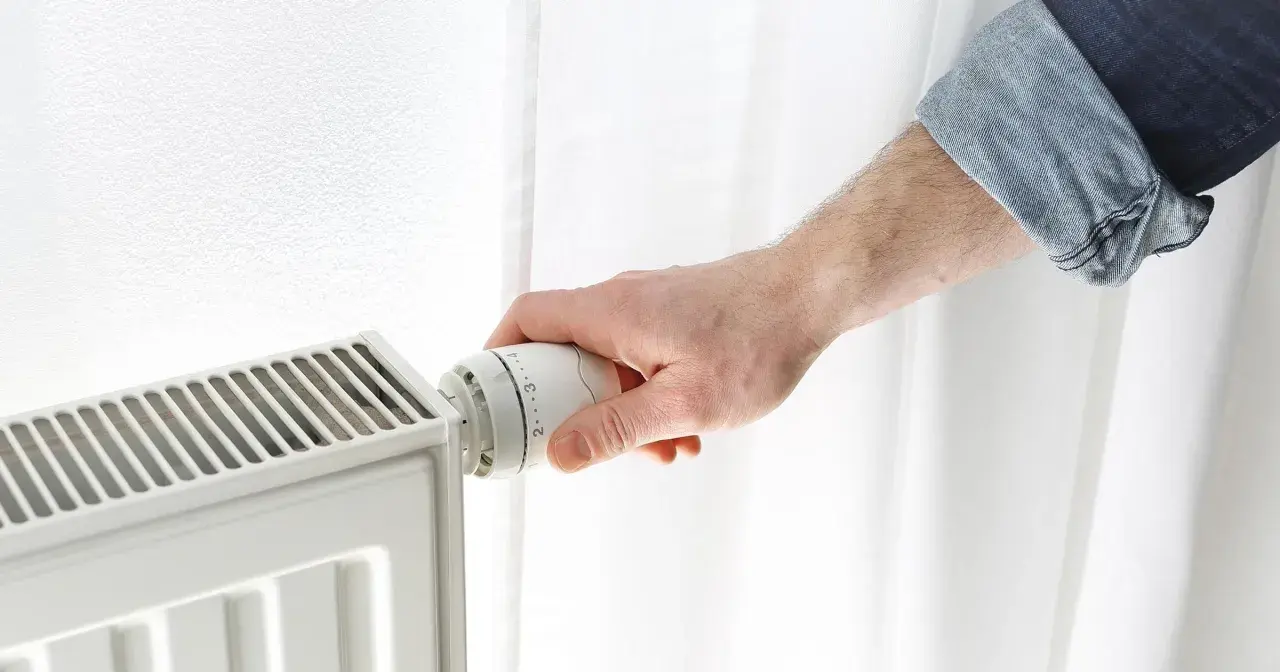 Dłoń reguluje termostat grzejnika, próbując obniżyć zbyt wysoką temperaturę w domu.