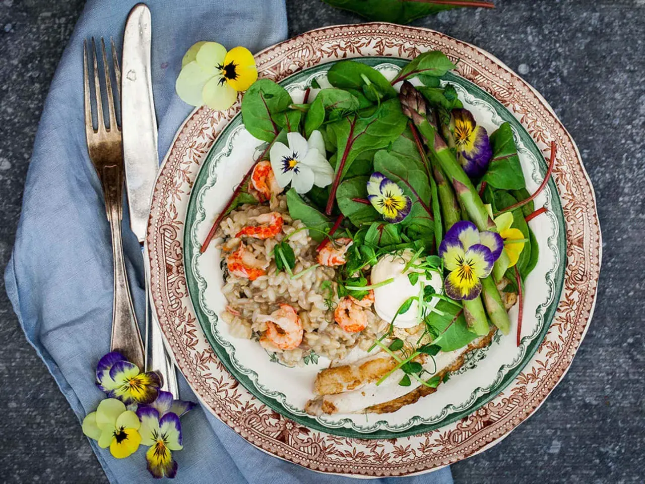 A plate of creamy risotto with shrimp, asparagus, greens, and edible flowers, served with a fork and knife.