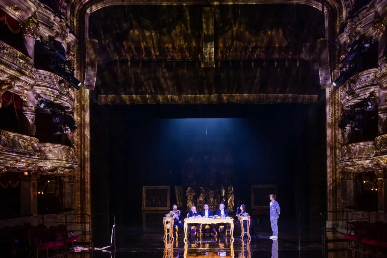 Na scenie teatru, przy złotym stole, siedzi pięć postaci. W tle widać bogato zdobione loże i kurtyny, tworząc atmosferę historycznego przedstawienia.
