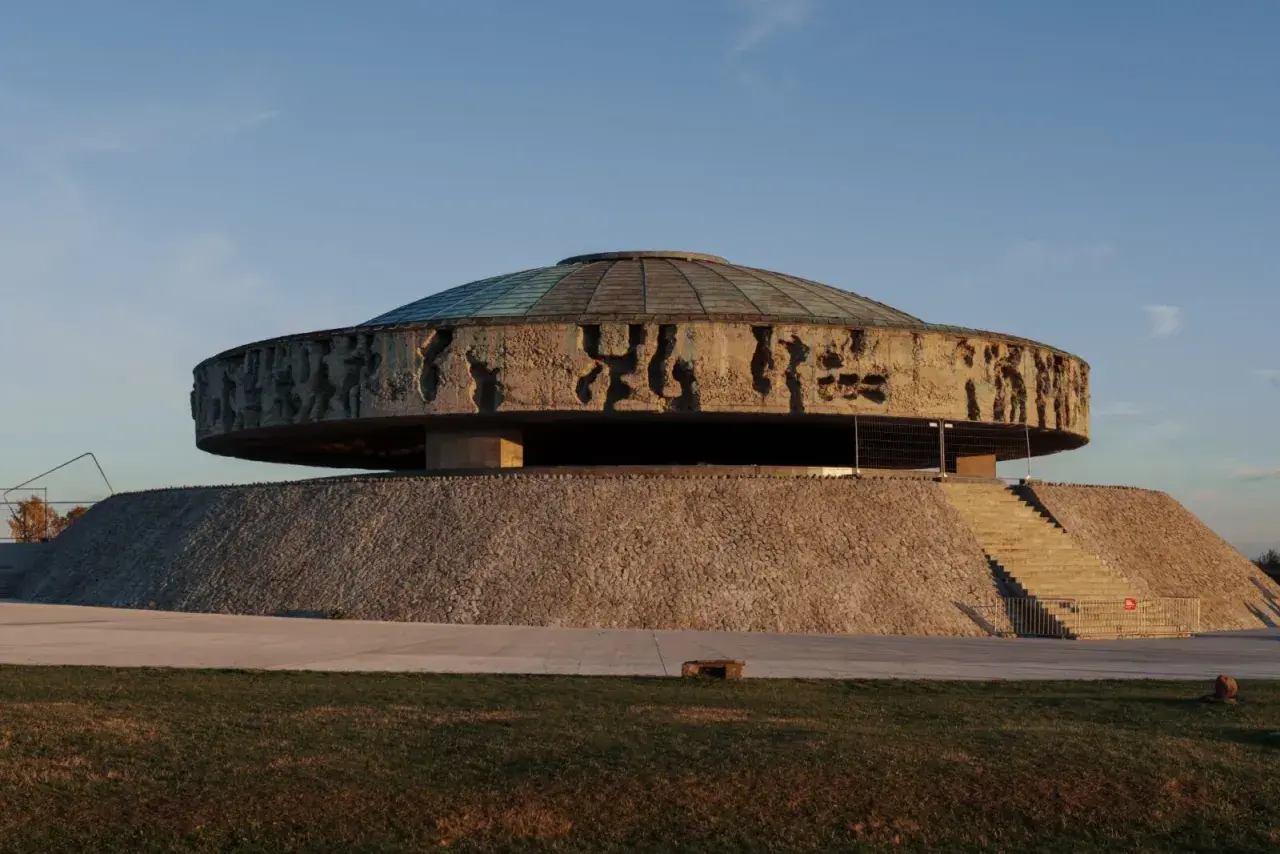 Monumentalne mauzoleum o okrągłej podstawie, z betonowym dyskiem ozdobionym płaskorzeźbami i szklanym dachem.