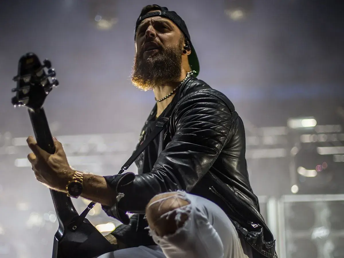 Charyzmatyczny gitarzysta z brodą podczas koncertu Bullet for My Valentine, w skórzanej kurtce i podartych spodniach, gra na gitarze.