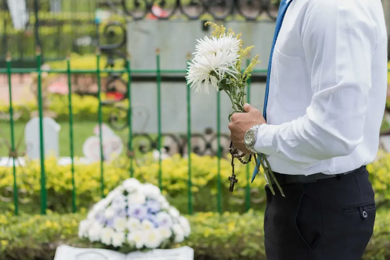Mężczyzna w białej koszuli i niebieskim krawacie trzyma bukiet białych kwiatów i różaniec. To jego urlop okolicznościowy śmierć bliskiej osoby.