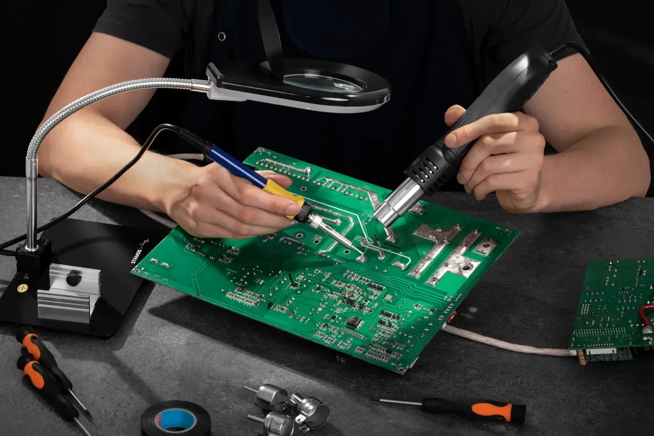 Ręce lutują płytę elektroniczną, używając lutownicy i gorącego powietrza. Widać też lupę, śrubokręty i taśmę izolacyjną – wszystko, co można lutować.