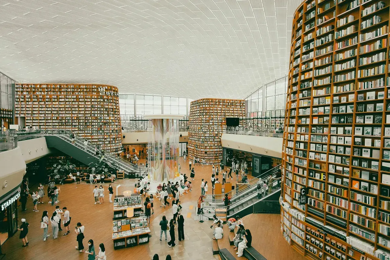 Wielopoziomowa biblioteka z półkami sięgającymi sufitu, gdzie każdy **bibliotekarz** marzyłby o pracy. Ludzie spacerują, czytają i odpoczywają wśród książek.