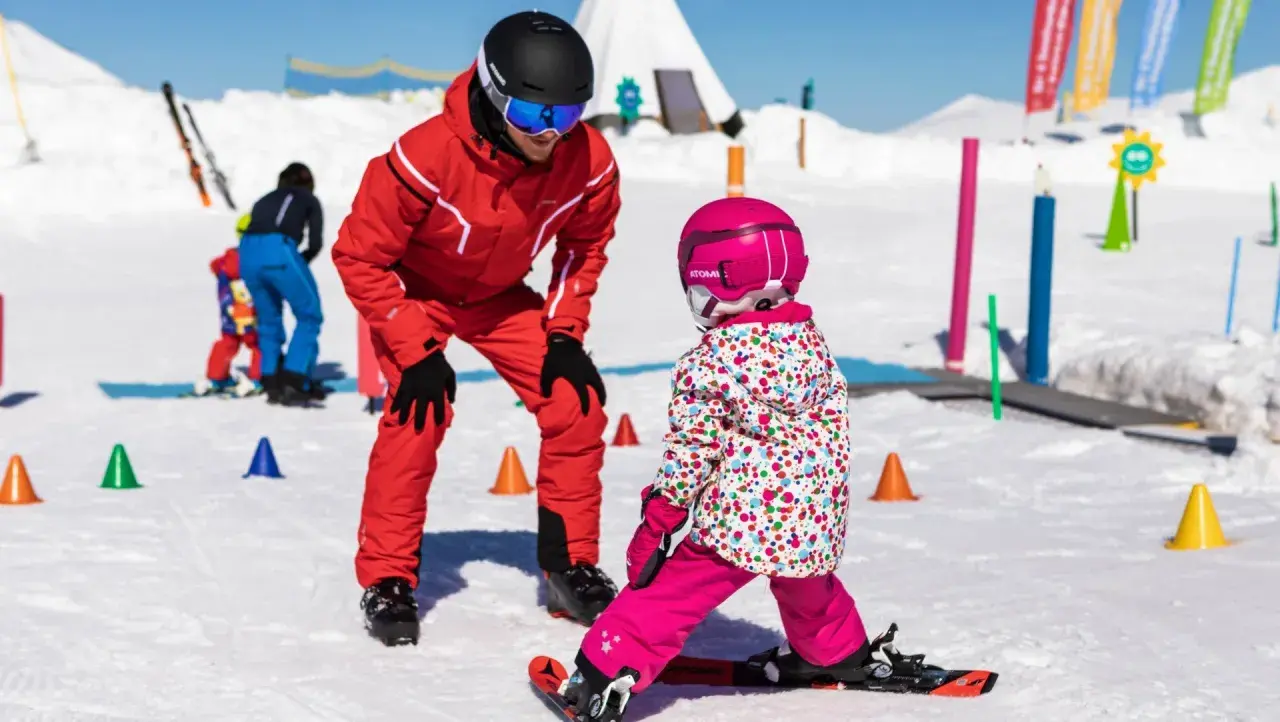 Maestro di sci insegna a bambini a sciare su pista innevata, con coni colorati e bandierine sullo sfondo.