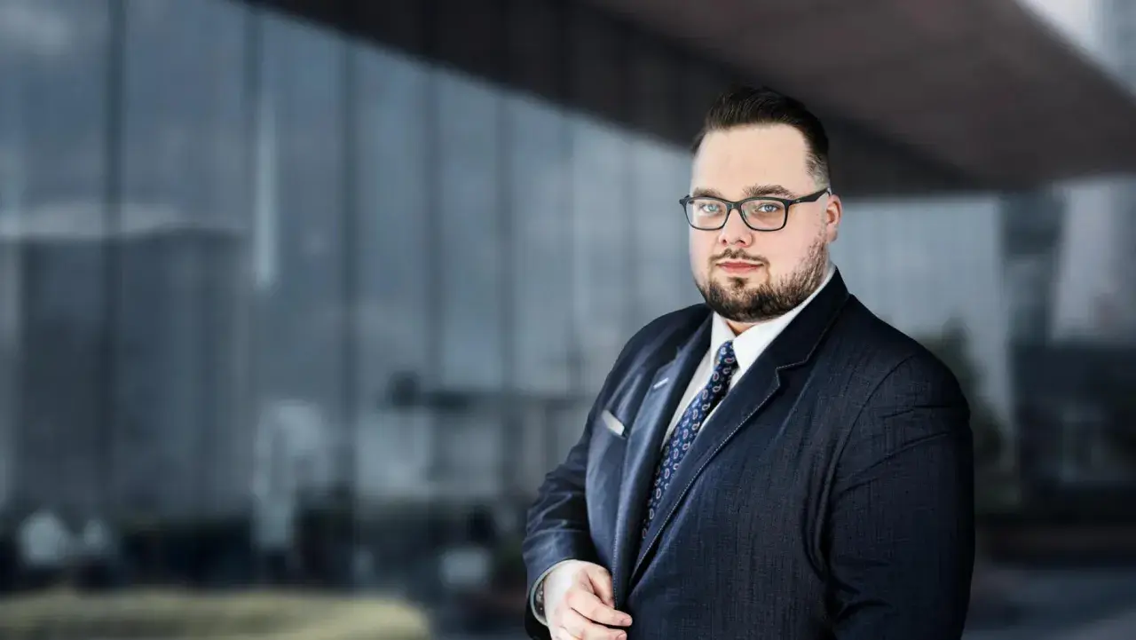 Mężczyzna w garniturze i okularach na tle nowoczesnego budynku.