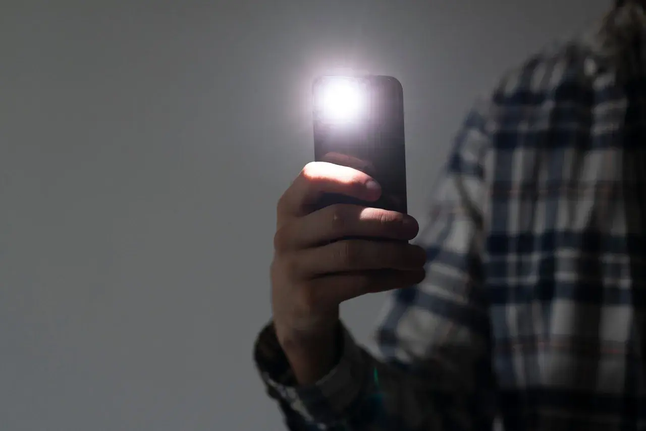 Dłoń trzyma telefon, którego latarka w telefonie rozświetla ciemność.