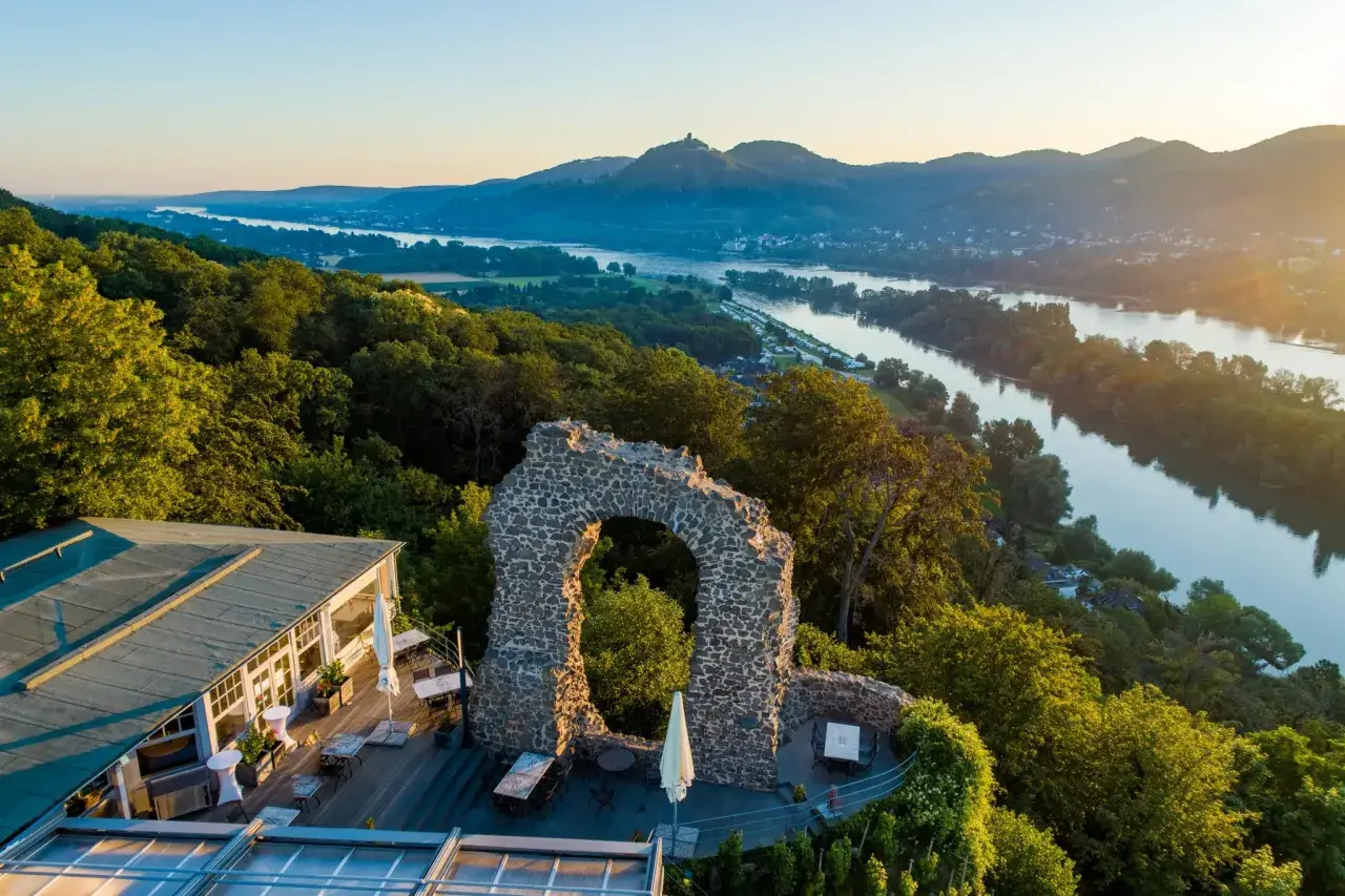 Der Rolandbogen thront über dem Rhein. Ein Restaurant mit Terrasse lädt zum Verweilen ein, während im Hintergrund Berge und ein Fluss im Abendlicht glänzen.