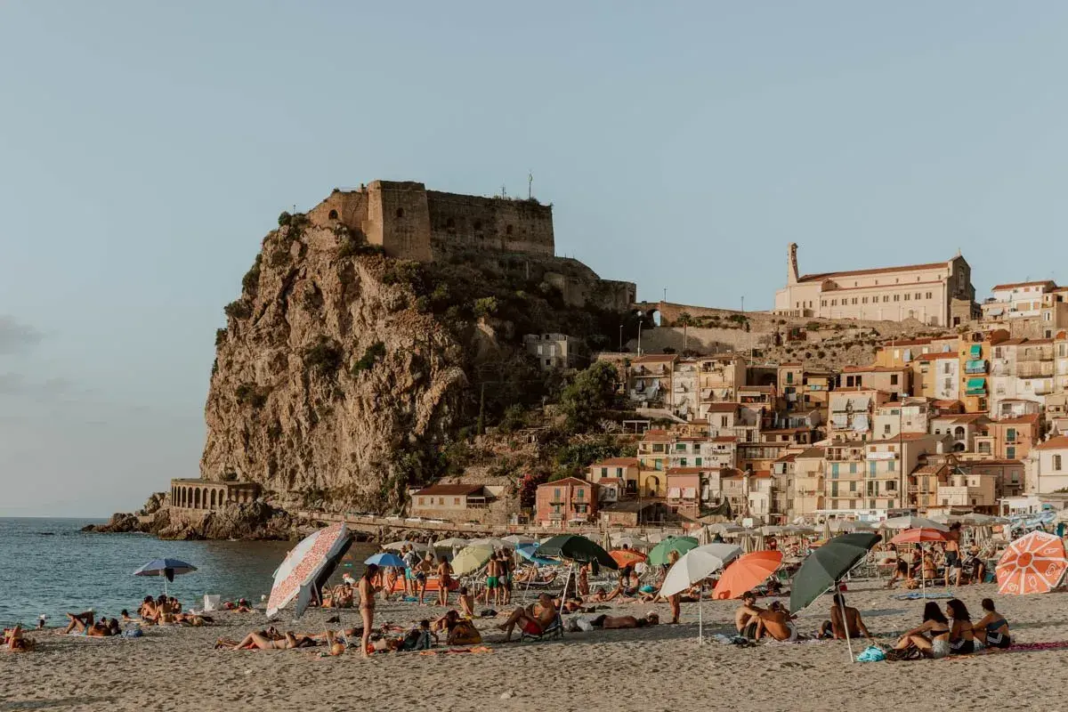 A sunny beach with people relaxing under umbrellas, overlooking a cliffside town with a castle. This view is a perfect destination for those wondering how to get to Calabria, Italy.