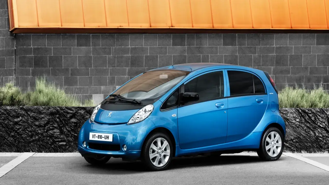 En blå Peugeot iOn står parkerad framför en grå tegelvägg med en orange takkant.