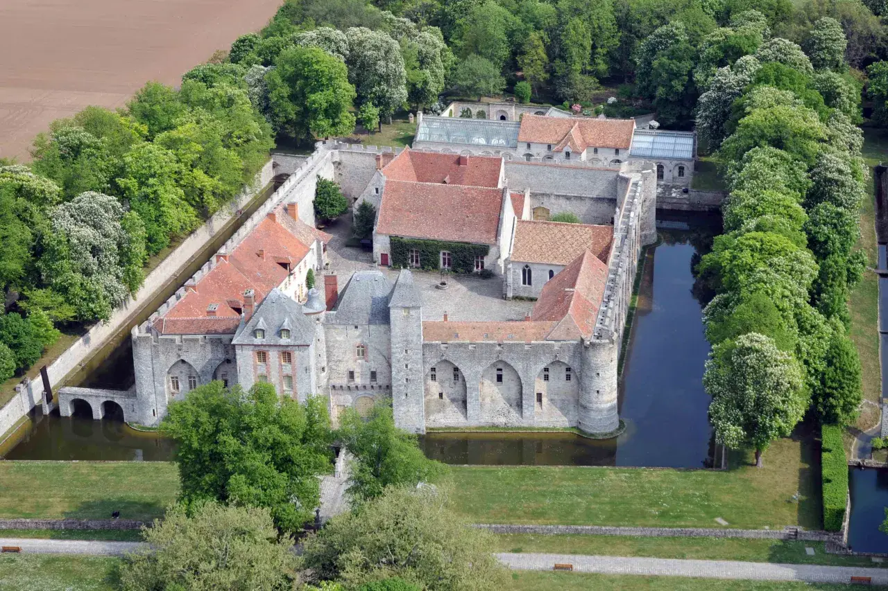 Vue aérienne du **château de Farcheville**, entouré de douves et de verdure. Ses toits de tuiles rouges contrastent avec les murs de pierre grise.