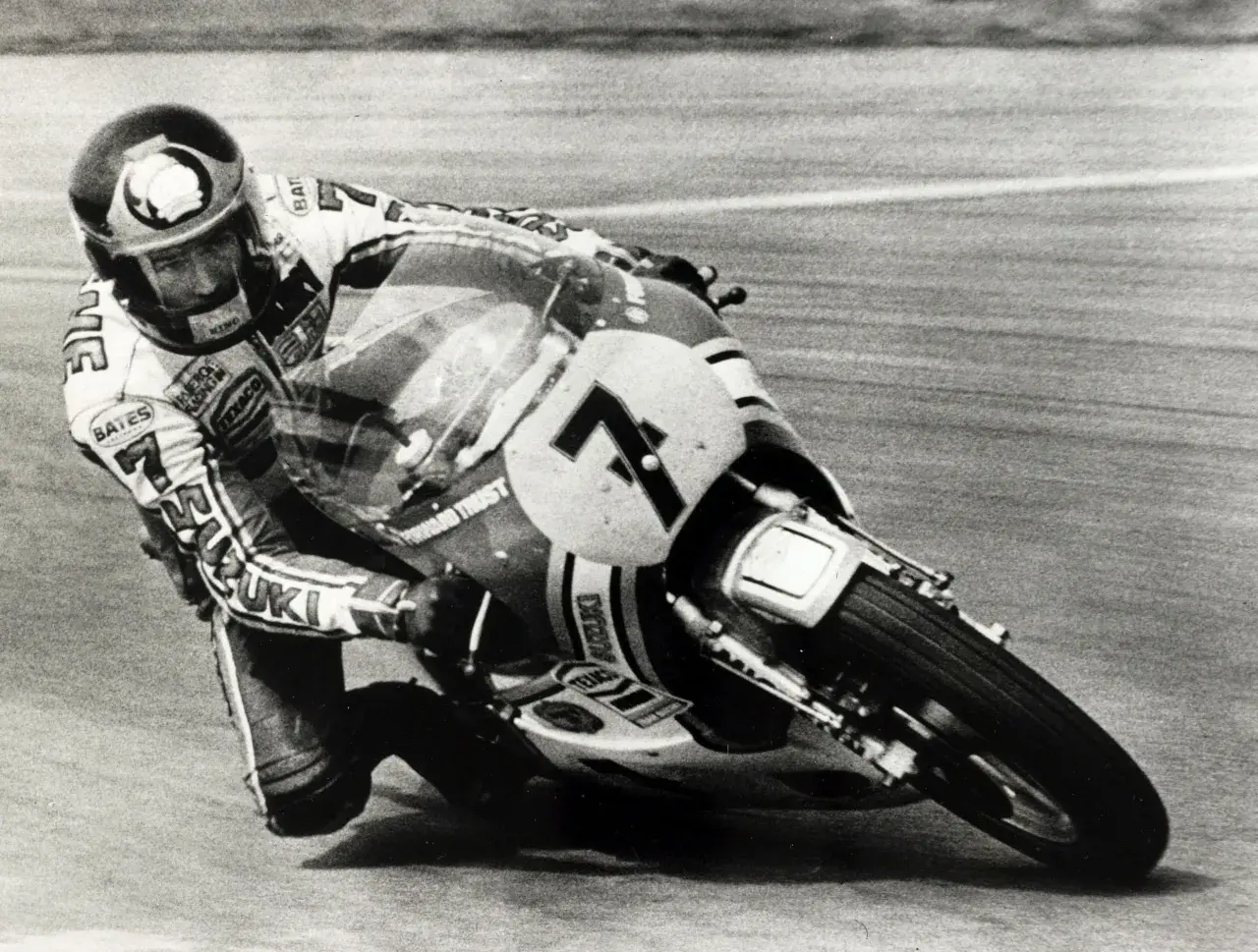 Wyścigowy motocykl Suzuki z numerem 7 pochylony w zakręcie, z zawodnikiem w kasku. To fragment historii motocykla.