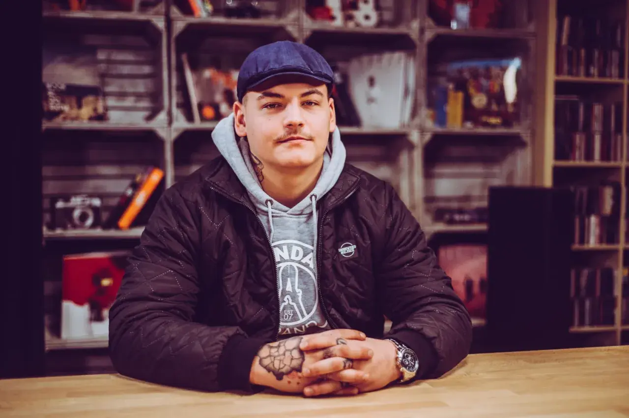 Młody mężczyzna w czapce i kurtce, z tatuażami na rękach, siedzi przy drewnianym stole. Wygląda jakby przeżywał swój **frosti wiek**, pełen pasji i determinacji.