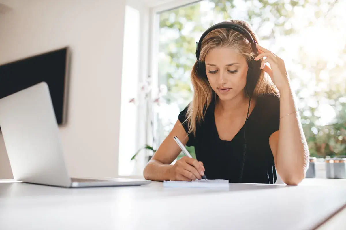 Mujer con auriculares escribe en un cuaderno, con luz natural entrando por la ventana. La luz suave es ideal, preguntándose cuál es la mejor luz para estudiar.