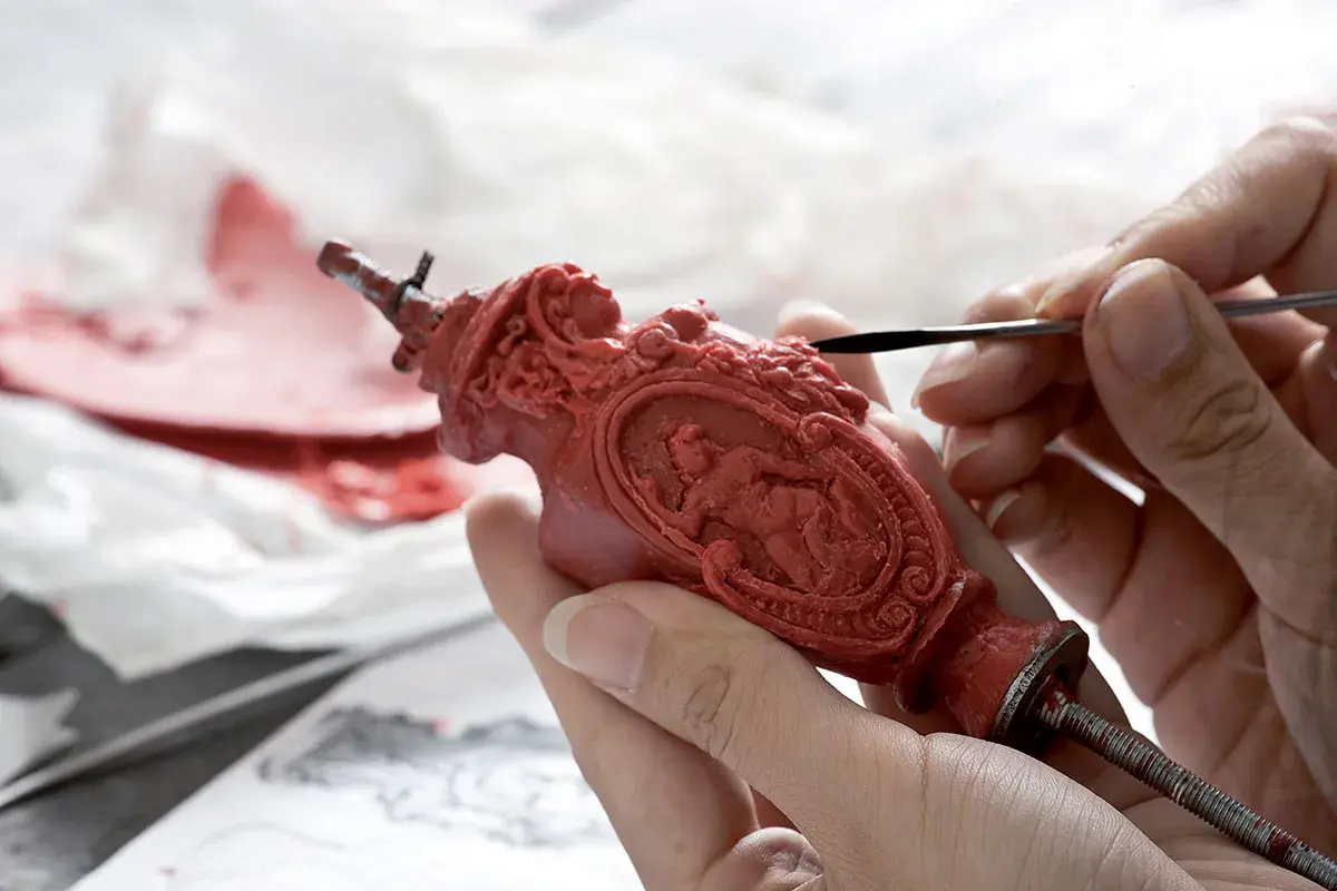 Mani esperte rifiniscono un oggetto in cera rossa, un'opera d'arte che ricorda la **fondazione Cologni**. Dettagli intricati prendono forma sotto gli strumenti.