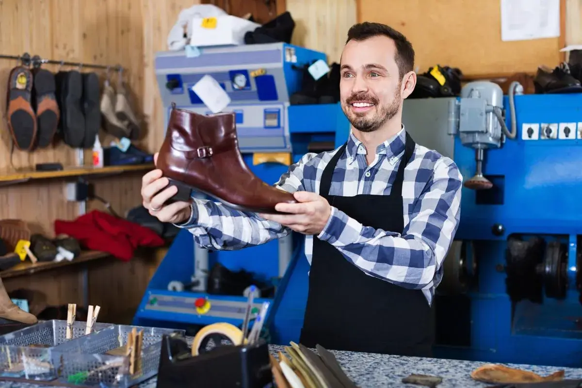 Artisan souriant tenant une botte en cuir marron, prêt pour la réparation de chaussure. L'atelier est bien équipé.