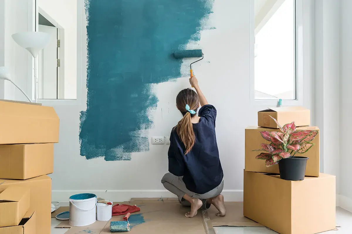 Mujer pintando una pared de azul, rodeada de cajas. Es la mejor época para pintar la casa y darle un nuevo aire.