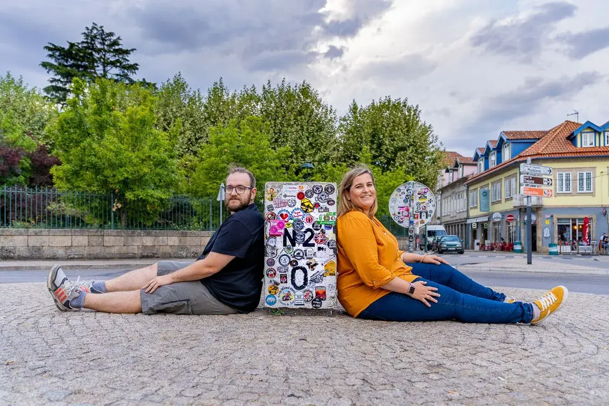 Pareja sentada de espaldas en un poste con pegatinas, celebrando su viaje por la Estrada Nacional 2 en Portugal.