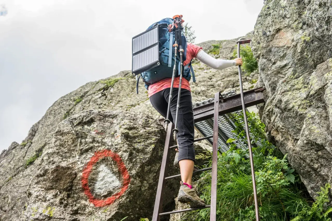 A hiker ascends metal ladders on the Tour du Mont Blanc, using trekking poles and a solar panel on their backpack.