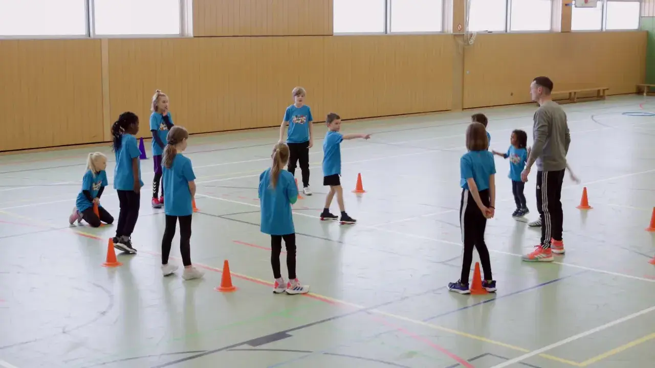 Kinder in blauen T-Shirts üben in der Turnhalle, während ein Trainer Anweisungen gibt. Tip tap, die Füße bewegen sich im Takt.