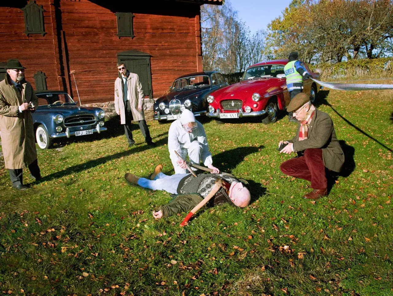 En brottsplats med en död man på marken, omgiven av gamla bilar, inklusive en British Leyland. Polis och utredare arbetar.