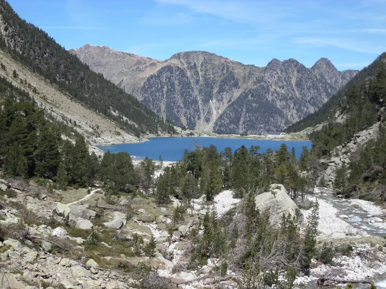 Lago azul rodeado de montañas y pinos, con un pequeño refugio en la orilla. El refugio Oulettes de Gaube es un destino popular para excursionistas.