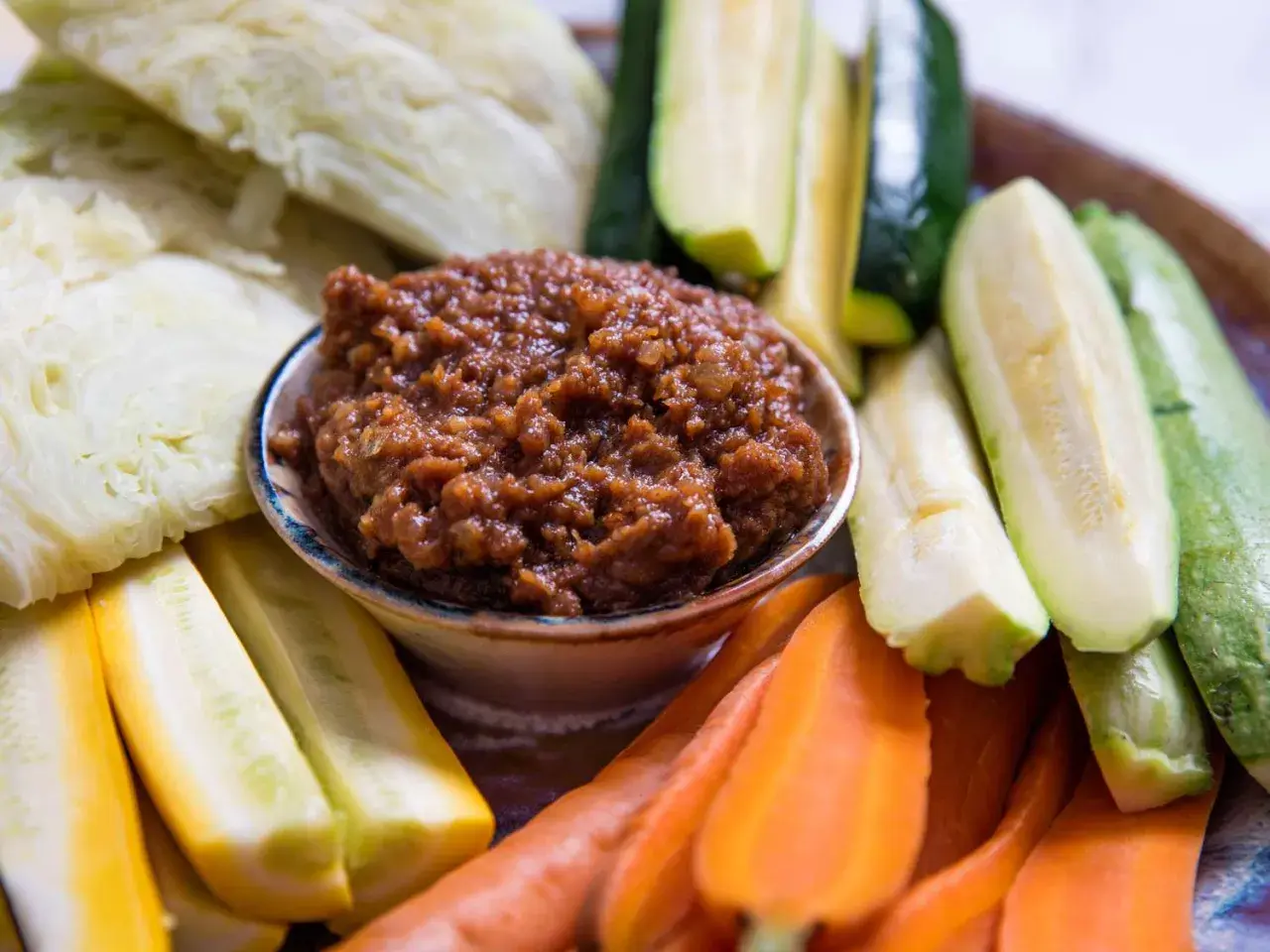 A bowl of savory miso dip is surrounded by fresh, colorful crudités: sliced zucchini, yellow squash, carrots, and cabbage wedges, ready for dipping.