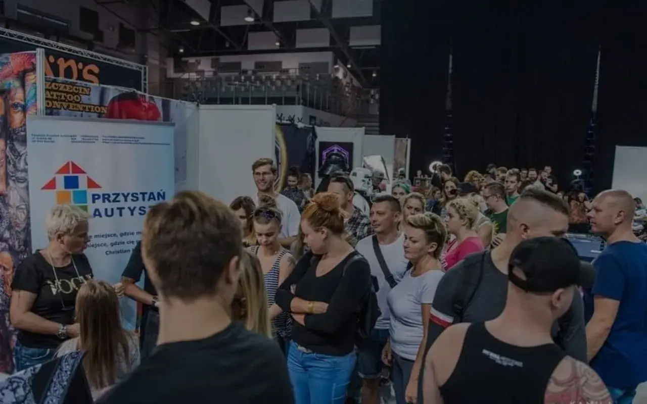 Ludzie na konwencie tatuażu, gdzie widoczny jest symbol autyzmu na banerze "Przystań Autys".