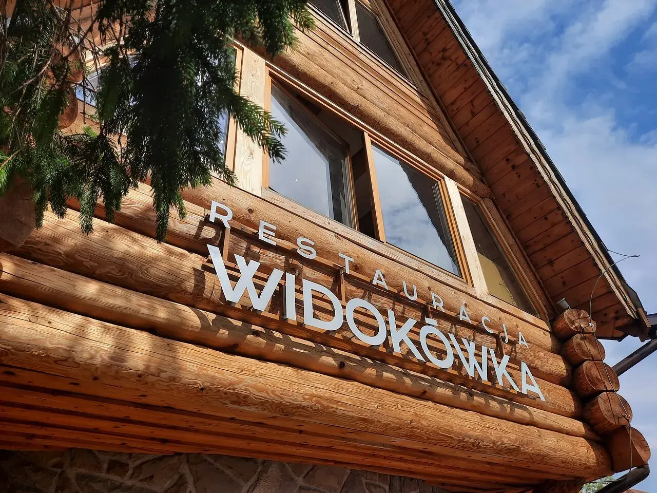 Drewniana restauracja Widokówka z białym napisem. Gałązka świerku zwisa z lewej strony.
