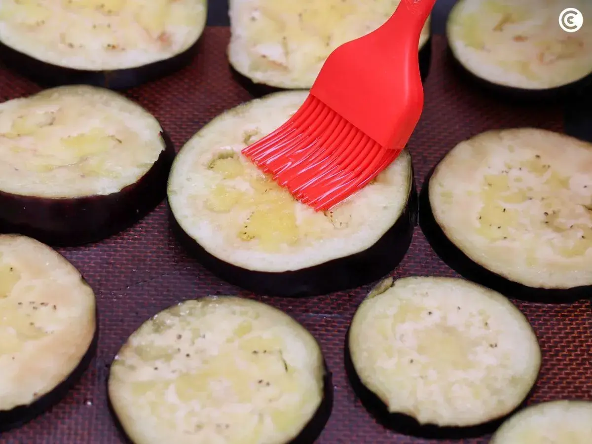 Rodajas de berenjenas listas para ser horneadas, untadas con aceite y especias con un pincel rojo.