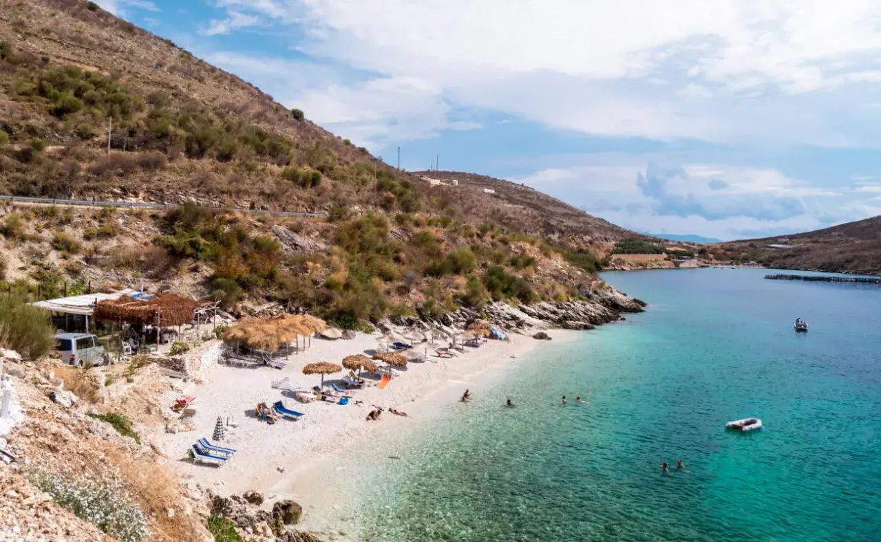 Słoneczna plaża w Porto Palermo z turkusową wodą, leżakami i łódkami.