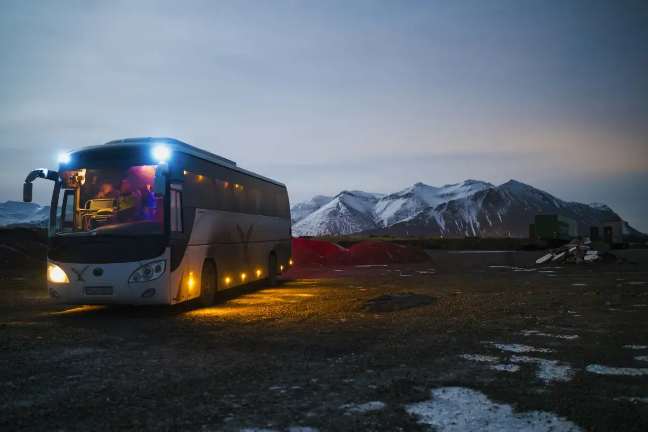 Autobus z zapalonymi światłami na tle ośnieżonych gór o zmierzchu.