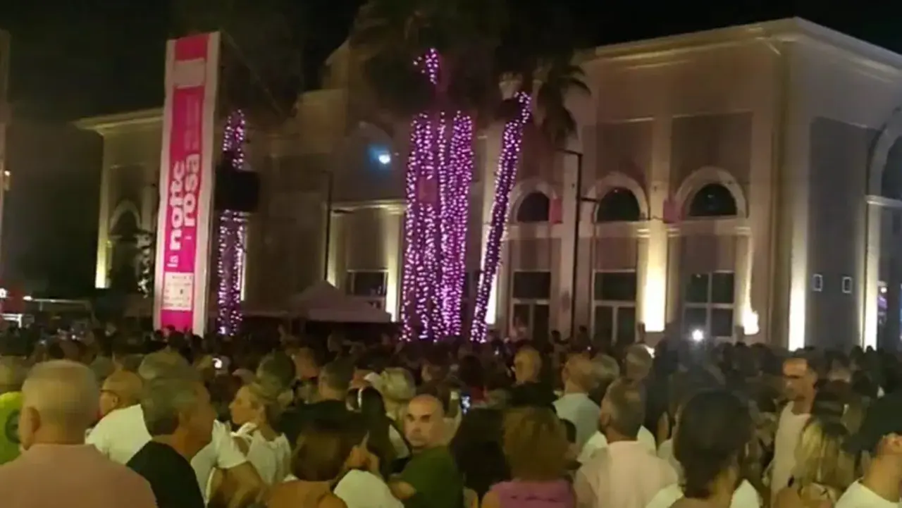 Folla festante a Porto Sant'Elpidio per la Notte Rosa, con palme illuminate di rosa e un grande striscione.